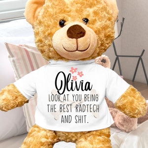 May include: A large, brown teddy bear wearing a white t-shirt with the text "Olivia LOOK AT YOU BEING THE BEST RADTECH AND SHIT." printed in black. The bear is sitting on a white and pink striped bedspread. The bear is 22 inches tall.