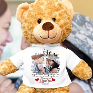 May include: A plush teddy bear with light brown fur. The bear wears a white t-shirt with a photo and text that reads "MY DEAR Olivia" and "I Love You". The bear is a sentimental gift.