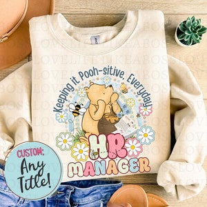 May include: A beige sweatshirt with a Winnie the Pooh illustration and the text "Keeping it Pooh-sitive, Everyday". The illustration shows Winnie the Pooh holding a book and sitting on a honey pot. The text "HR Manager" is below the illustration. The sweatshirt has a custom option for any title.