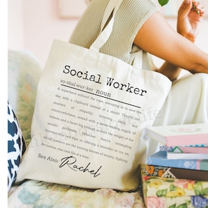 May include: Cream-colored tote bag with the words "Social Worker" printed in black. The bag features a definition of a social worker and the name "Rachel" in script. The bag is held by a person.