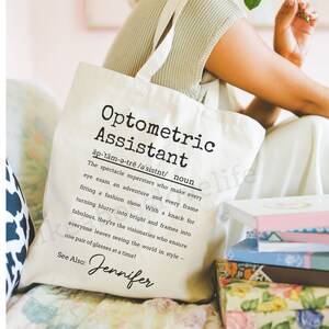 May include: A white canvas tote bag with a black print that defines "Optometric Assistant" as "The spectacle superstars who make every eye exam an adventure and every frame fitting a fashion show." The text also includes "See Also: Jennifer".