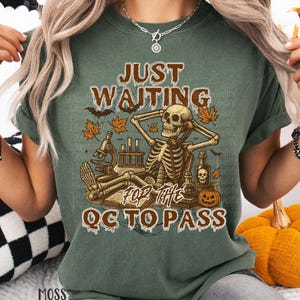 May include: A green t-shirt with a graphic featuring a skeleton relaxing with lab equipment and the text "JUST WAITING FOR THE QC TO PASS". The design includes fall leaves and a pumpkin, perfect for Halloween.