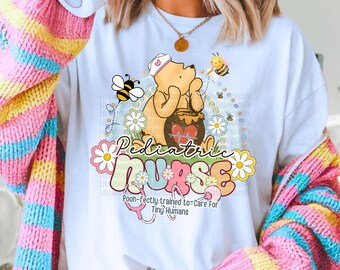 May include: White t-shirt with a graphic featuring Winnie the Pooh as a pediatric nurse, surrounded by bees and flowers. The text reads "Pediatric Nurse" and "Pooh-fectly trained to care for tiny humans."