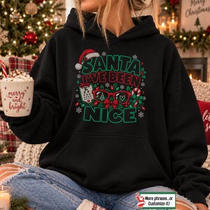 May include: A black hooded sweatshirt featuring a graphic print with the text "SANTA I'VE BEEN NICE" in green and red. The design includes a Santa hat, red eyeglasses, a vision chart, a candy cane, and holly leaves surrounded by snowflakes.