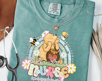 May include: Light green t-shirt with a graphic featuring Winnie the Pooh as a pediatric nurse, bees, and daisies. The text reads "Pediatric Nurse" and "Pooh-fectly trained to care for tiny humans."