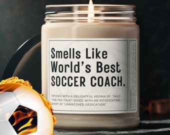 May include: A white candle with a black label that reads "Smells Like World's Best Soccer Coach." The label also includes the text "Infused with a delightful aroma of "Half-Time-Pep-Talk!" mixed with an intoxicating whiff of "Unmatched-Dedication"". A soccer ball with flames is in the foreground.