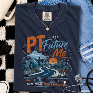 May include: A navy blue t-shirt featuring a graphic print with the text PT FOR Future Me. The design includes a winding road leading toward a sunset over mountains, with silhouettes of people exercising, the name Sarah, and the words Strength, Mobility, Resilience, Wellness, Move Today, Thrive Tomorrow.