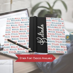 May include: A black notebook with the name "Belinda" written in white lettering on the front cover. The notebook is open to a page with a variety of text in red, blue, and white. The text is a code or a pattern of letters and numbers.