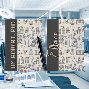 May include: Two black and white science themed notebooks with the names "Jim Robert, PhD" and "Elaine" on the covers. The notebooks are sitting on a table in a laboratory setting.