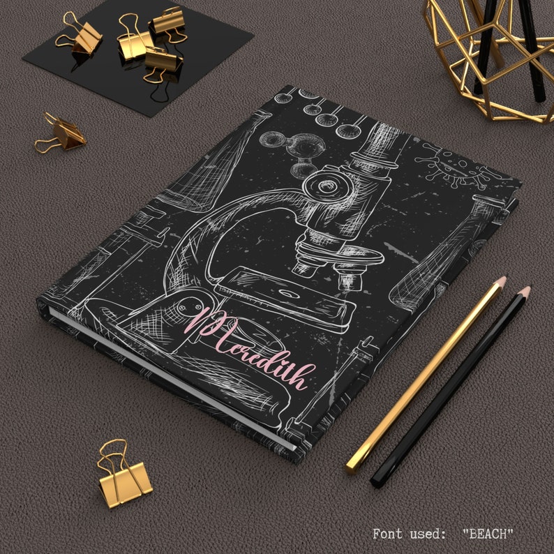 Peut inclure: Un carnet noir avec un microscope blanc et d'autres illustrations scientifiques. Le nom "Meredith" est &eacute;crit en rose sur la couverture.