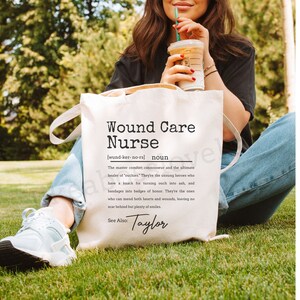 May include: White tote bag with black text that reads "Wound Care Nurse (wund-ker-no-rs) noun The master comfort connoisseur and the ultimate healer of 'ouches'. They're the unsung heroes who have a knack for turning ouch into ah, and bandages into badges of honor. They're the ones who can mend both hearts and wounds, leaving no scar behind but plenty of smiles. See Also: Taylor"