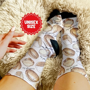 May include: White socks with a repeating pattern of sourdough bread illustrations. The socks have black heels and toes. The text "UNISEX SIZE" is in a red starburst shape.
