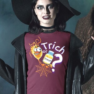 以下が含まれることがあります： ハロウィーンをテーマにしたバーガンディ色のTシャツ。デザインは、笑顔の漫画の細菌、"Treat"と書かれた薬瓶、クモの巣、"Trick or ?"の文字が特徴です。黒い服を着た人が着用しています。