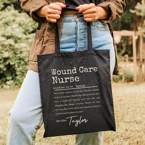 May include: Black tote bag with white text that reads "Wound Care Nurse [wund-ker-no-rs] noun The master comfort connoisseur and the ultimate healer of 'outhies'. They're the unsung heroes who have a knack for turning ouch into ash, and bandages into badges of honor. They're the ones who can mend both hearts and wounds, leaving no scar behind but plenty of smiles. See Also: Taylor"