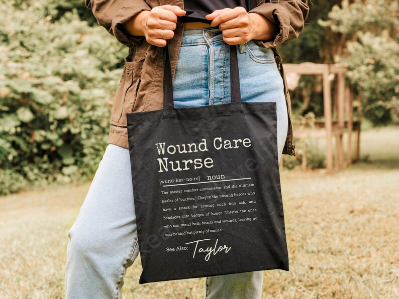 May include: Black tote bag with white text that reads "Wound Care Nurse [wund-ker-no-rs] noun The master comfort connoisseur and the ultimate healer of 'outhies'. They're the unsung heroes who have a knack for turning ouch into ash, and bandages into badges of honor. They're the ones who can mend both hearts and wounds, leaving no scar behind but plenty of smiles. See Also: Taylor"