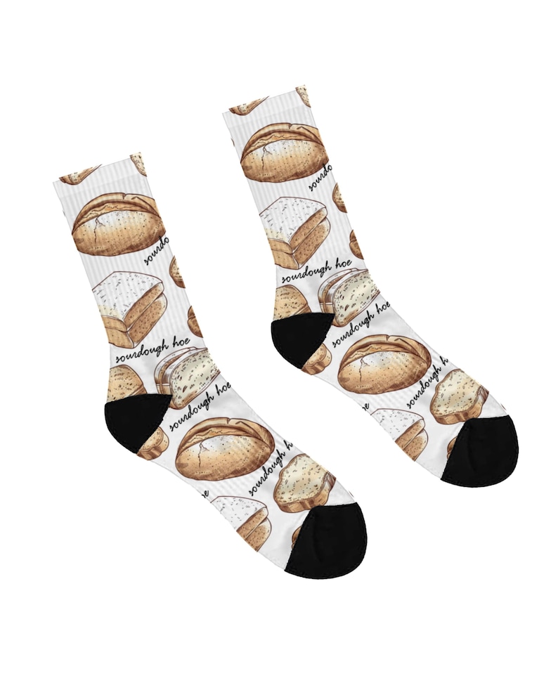May include: A pair of white socks with a repeating pattern of watercolor-style sourdough bread loaves and slices. The socks have black heels and toes, and the words "sourdough hoe" are printed on the socks.
