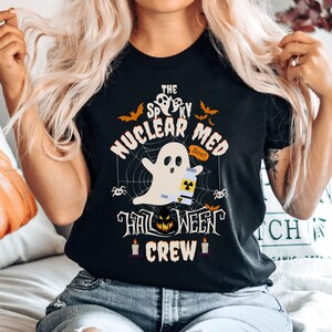 May include: Black t-shirt with a Halloween-themed graphic. The design features a ghost holding a vial, surrounded by a spider web, bats, and the text "The Spooky Nuclear Med Halloween Crew".
