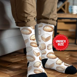 May include: White socks with a repeating pattern of various bread loaves and slices in shades of brown. The socks have black heels and toes, and a red "UNISEX SIZE" sticker is visible.