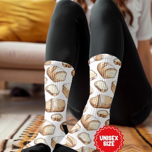 May include: White socks with a repeating pattern of illustrated bread loaves and slices. The socks have black toes and heels. A red sticker with the words "UNISEX SIZE" is visible.