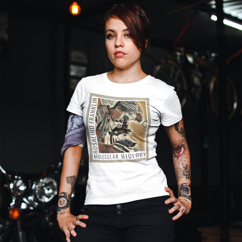 May include: White t-shirt with a vintage-style illustration of Rosalind Franklin, a scientist, looking through a microscope. The text "Rosalind Franklin" and "Molecular Biology" is printed on the shirt.