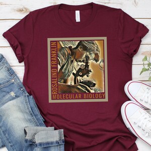 May include: A maroon t-shirt with a vintage-style illustration of Rosalind Franklin looking through a microscope. The text "Rosalind Franklin" and "Molecular Biology" is printed on the shirt.