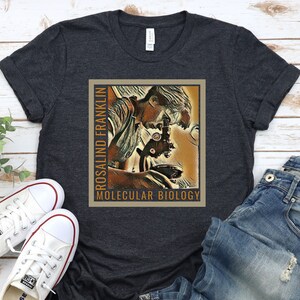 May include: A dark grey t-shirt with a vintage-style illustration of Rosalind Franklin looking through a microscope. The text "Rosalind Franklin" and "Molecular Biology" is printed on the shirt.