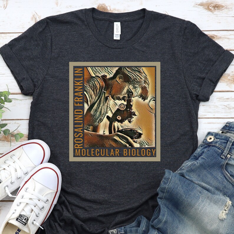 May include: A dark grey t-shirt with a vintage-style illustration of Rosalind Franklin looking through a microscope. The text "Rosalind Franklin" and "Molecular Biology" is printed on the shirt.