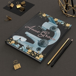May include: A black notebook with a watercolor illustration of a microscope and floral accents. The notebook has the name "Anne Grace" written in pink script.