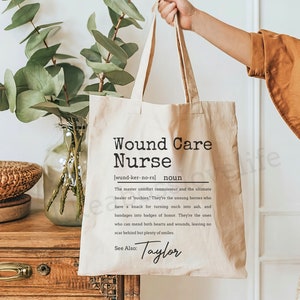 May include: A beige tote bag with a black and white design that reads "Wound Care Nurse (wund-ker-no-rs) noun The master comfort connoisseur and the ultimate healer of 'outhies.' They're the unsung heroes who have a knack for turning ouch into ash, and bandages into badges of honor. They're the ones who can mend both hearts and wounds, leaving no scar behind but plenty of smiles See Also: Taylor"