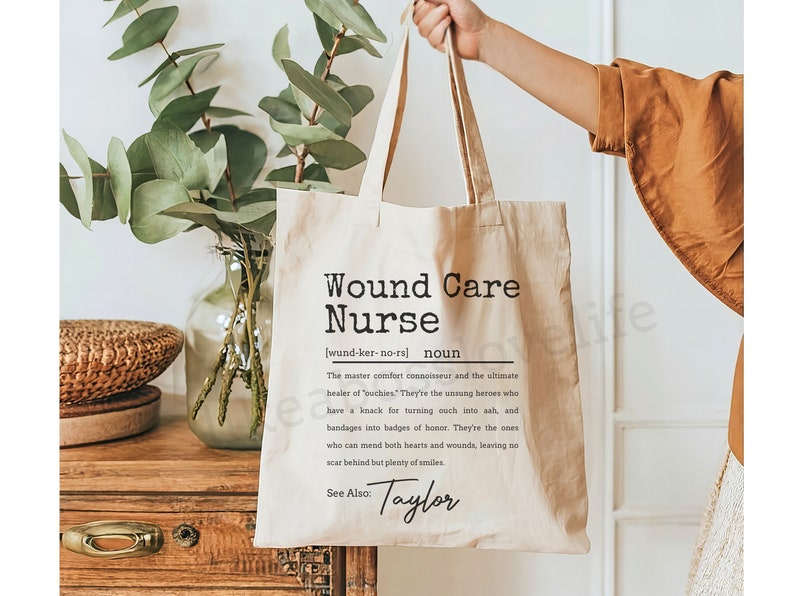 May include: A beige tote bag with a black and white design that reads "Wound Care Nurse (wund-ker-no-rs) noun The master comfort connoisseur and the ultimate healer of 'outhies.' They're the unsung heroes who have a knack for turning ouch into ash, and bandages into badges of honor. They're the ones who can mend both hearts and wounds, leaving no scar behind but plenty of smiles See Also: Taylor"