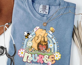 May include: Blue jean colored t-shirt with a graphic of Winnie the Pooh as a nurse, bees, and the words "Pediatric Nurse." The shirt has a crew neck and short sleeves. A stethoscope and necklace are also visible.