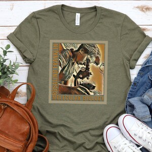 May include: A green t-shirt with a vintage-style illustration of Rosalind Franklin looking through a microscope. The text "Rosalind Franklin" and "Molecular Biology" is printed on the shirt.