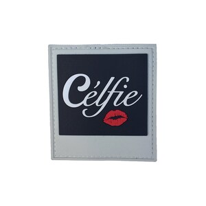 以下が含まれることがあります： 白い文字で「Céffie」と赤い唇のプリントが施された、黒い中心部を持つ、薄い灰色の正方形のパッチ。パッチには白い縁とステッチされたエッジがあります。