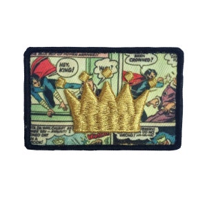 Può includere: Un patch ricamato con una corona dorata su sfondo di fumetto. Lo sfondo presenta un supereroe con un mantello rosso e le parole "Hey, King!" e "Been Crowned?"