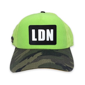 Può includere: Un cappello da baseball verde neon con una toppa nera che dice "LDN" in lettere bianche. Il cappello ha un motivo mimetico sul visore.