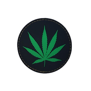 May include: A round, black patch with a green marijuana leaf design. The patch has a yellow stitched border.