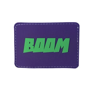 Peut inclure: Patch violet avec des coutures blanches autour des bords. Le patch a le mot "BOOM" en lettres vertes.