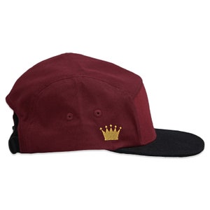 May include: A maroon 5-panel cap with a black brim and a gold crown embroidered on the front.