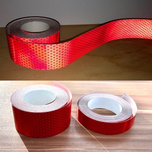 May include: Rolls of red reflective tape with a triangular pattern. The tape is designed to reflect light, enhancing visibility. The rolls are on a wooden surface, with one roll partially unrolled, displaying the tape's texture and color.