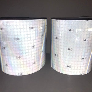 Puede incluir: Dos rollos de material reflectante plateado con un patrón repetitivo de pequeños cuadrados negros. El material es iridiscente y tiene un ligero efecto arcoíris.
