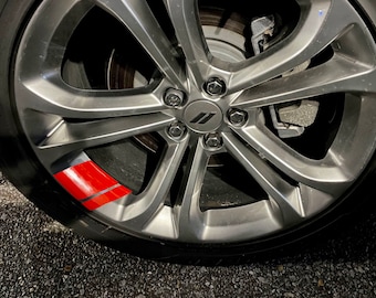 Puede incluir: Primer plano de una llanta de coche plateada con una calcomanía de rayas rojas y negras. La llanta tiene cinco radios y un logotipo de Dodge en el centro.