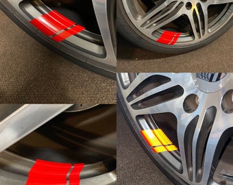Puede incluir: Primer plano de una llanta de coche plateada con rayas rojas. La rueda forma parte de un Porsche 911 Turbo. El neumático es negro y el fondo es una alfombra oscura.