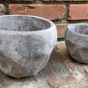 May include: Two gray concrete planters with a geometric design. The larger planter is in the foreground and the smaller planter is in the background.
