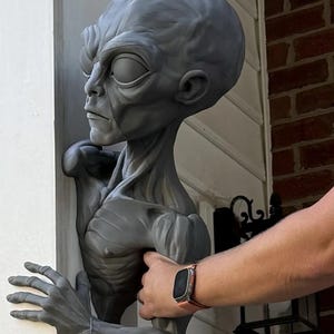 May include: A gray, sculpted bust of an extraterrestrial figure with a large, smooth cranium, prominent brow ridges, and deep-set eyes. The figure features a slender neck, defined chest musculature, and an extended arm with a hand reaching forward.