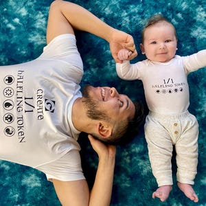 May include: A white t-shirt with the text "CREATE 1/1 HALFLING TOKEN" and a baby wearing a white onesie with the same text. The baby is wearing gray pants. Both are lying on a teal and blue textured surface.