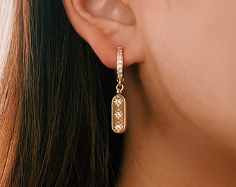 May include: Gold hoop earrings with a dangling charm featuring three small stars. The charm is a rectangular shape with a smooth, polished finish.