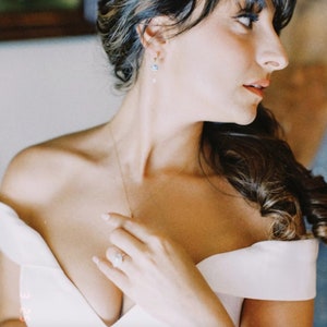 Op de afbeelding: Een bruid in een witte, off-shoulder jurk met een delicate ketting en oorbellen. Ze past met &eacute;&eacute;n hand de halslijn van haar jurk aan, terwijl haar andere hand op haar heup rust.
