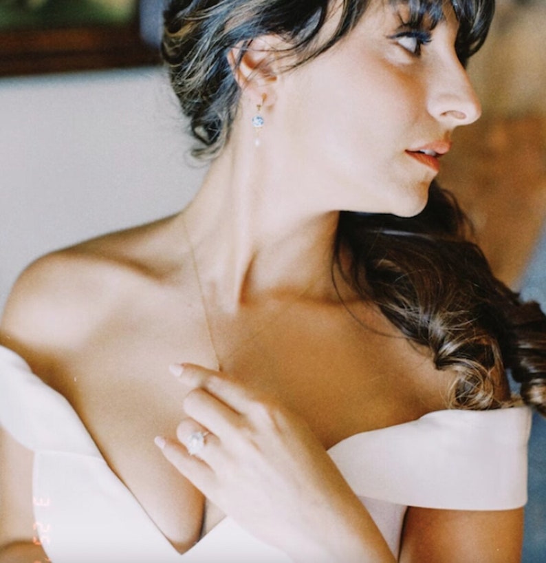 Op de afbeelding: Een bruid in een witte, off-shoulder jurk met een delicate ketting en oorbellen. Ze past met &eacute;&eacute;n hand de halslijn van haar jurk aan, terwijl haar andere hand op haar heup rust.