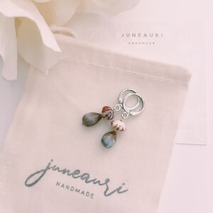 May include: A pair of silver hoop earrings with brown and white seed pod charms. The earrings are packaged in a white drawstring bag with the text "juneauri handmade" printed on it.