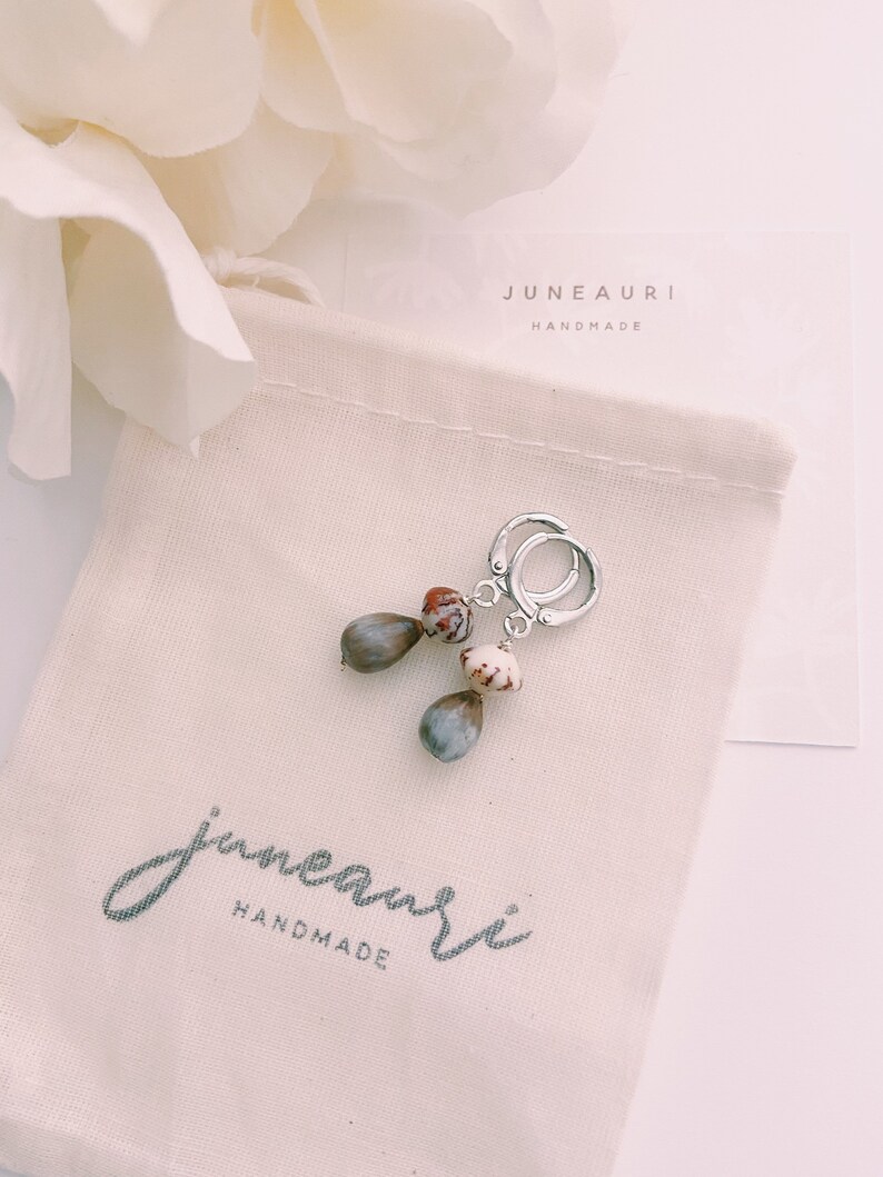 May include: A pair of silver hoop earrings with brown and white seed pod charms. The earrings are packaged in a white drawstring bag with the text "juneauri handmade" printed on it.
